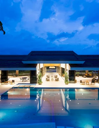 An illuminated resort or hotel pool area at night with lounge chairs and covered seating by the pool under a blue, clouded sky.