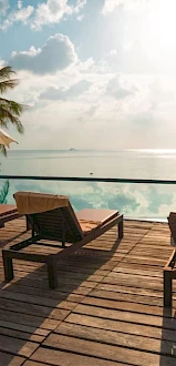 Lounge chairs and umbrellas by a pool with a view of the ocean, surrounded by palm trees and a sunny sky, creating a serene and relaxing scene.