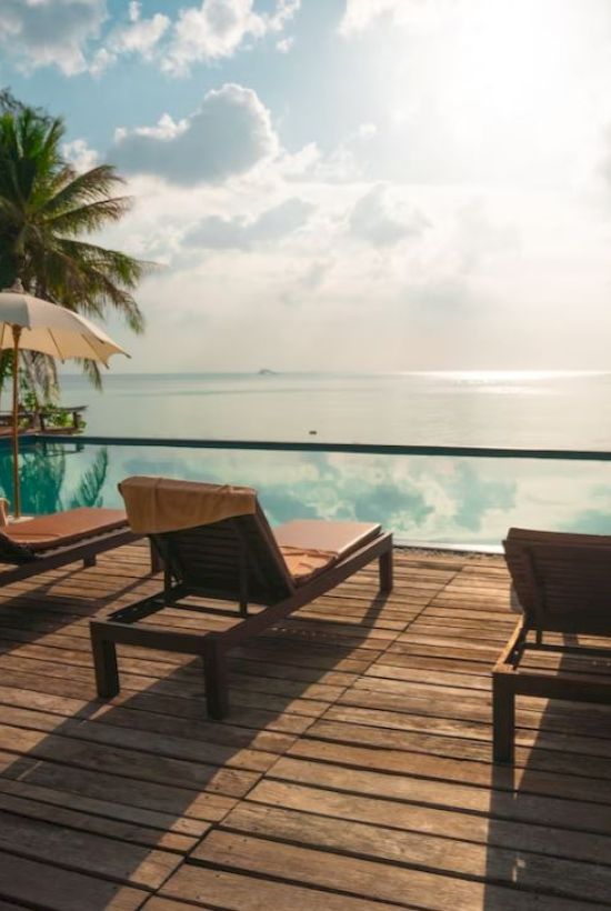The image shows a serene poolside with lounge chairs, umbrellas, palm trees, and a view of a calm sea under a partly cloudy sky, creating a tranquil scene.