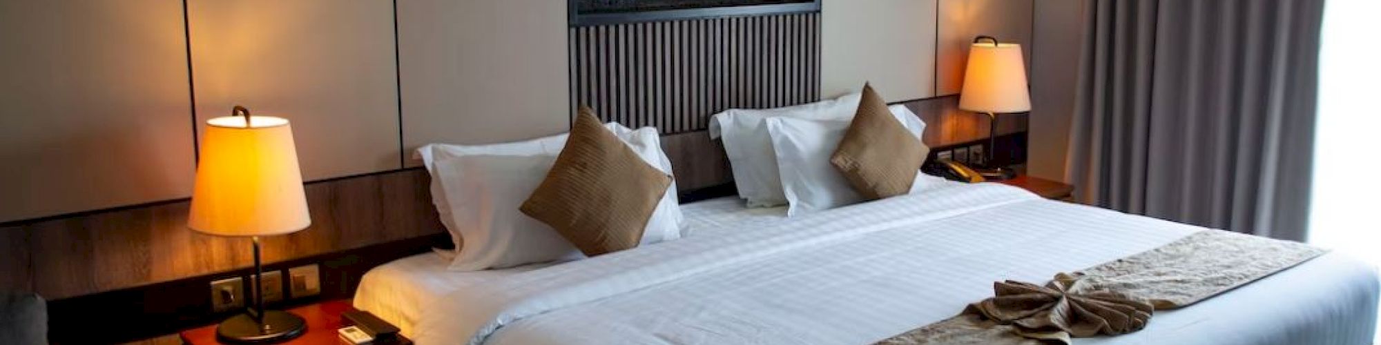 A cozy hotel room with a large bed, two bedside tables with lamps, decorative pillows, and a small table. There's a window with curtains.