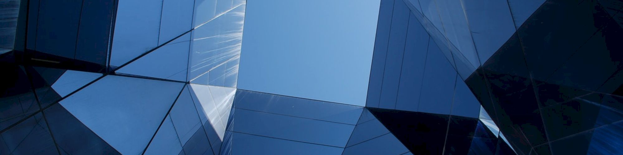 A view looking up through a geometric array of reflective glass panels with a clear blue sky visible in the center.