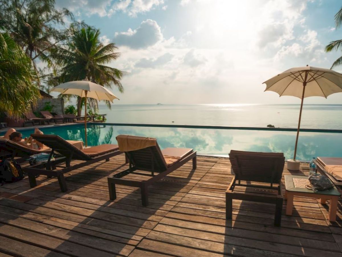 This image shows a serene poolside area with lounge chairs, umbrellas, palm trees, and a view of a calm sea under a partly cloudy sky.