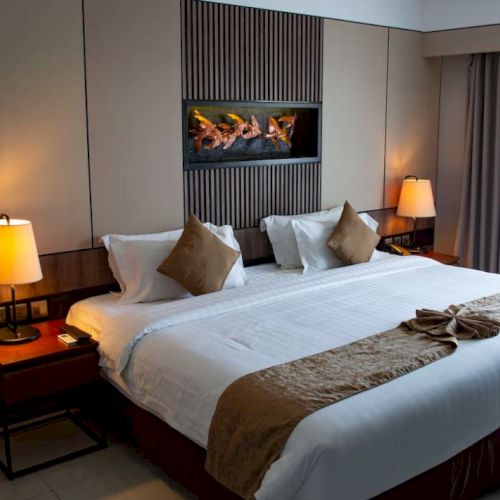 The image shows a cozy hotel room with a large bed, two bedside lamps, decorative pillows, and a wall-mounted decoration above the headboard.
