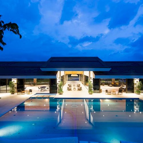 A luxurious outdoor pool area with a large, modern building behind it, illuminated under an evening sky.