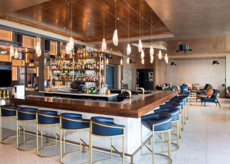 A modern bar with a wooden counter, blue and gold stools, hanging lights, and stocked shelves in a spacious, stylish setting.