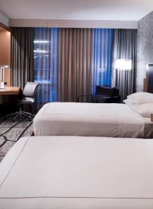 The image shows a modern hotel room with two double beds, a wall-mounted TV, a desk with a chair, and floor-to-ceiling windows with curtains.