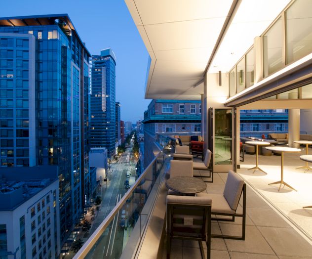 A modern balcony with seating overlooks a city street lined with tall buildings at dusk, featuring large windows and a sleek design.