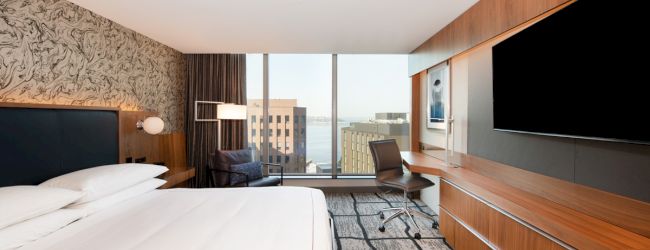 A modern hotel room with a large bed, a desk and chair, a wall-mounted TV, large windows offering a city view, and contemporary decor.