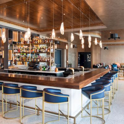 A modern bar with high chairs, a well-stocked liquor shelf, hanging pendant lights, and a sleek counter in a spacious, stylish setting.