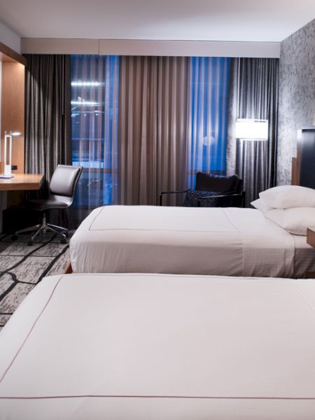 A modern hotel room with two beds, a TV on the wall, a work desk by the window, and minimalist decor featuring neutral colors and stylish furnishings.
