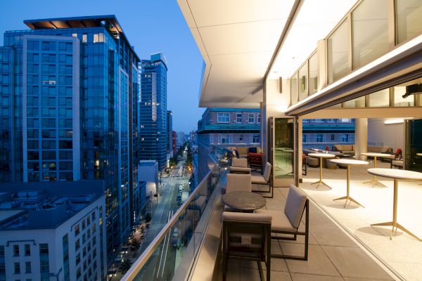 An illuminated balcony with tables and chairs overlooking a cityscape at dusk, featuring modern high-rise buildings and a vibrant street below.
