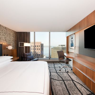 A modern hotel room featuring a large bed, a desk with a chair, a flat-screen TV, large windows with a city view, and a comfortable chair.