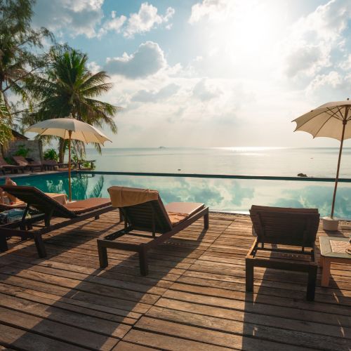 A serene beach scene featuring lounge chairs, umbrellas, and a pool by the water, with a stunning view of a tropical paradise under a bright sky.