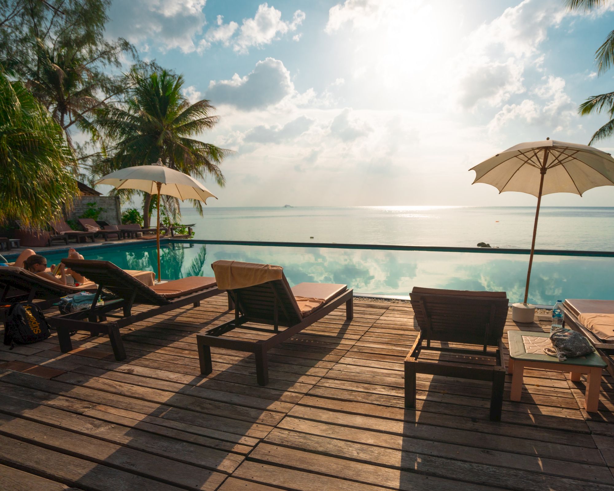 Lounge chairs and umbrellas by a pool, overlooking a serene ocean under a partly cloudy sky, surrounded by tropical greenery.