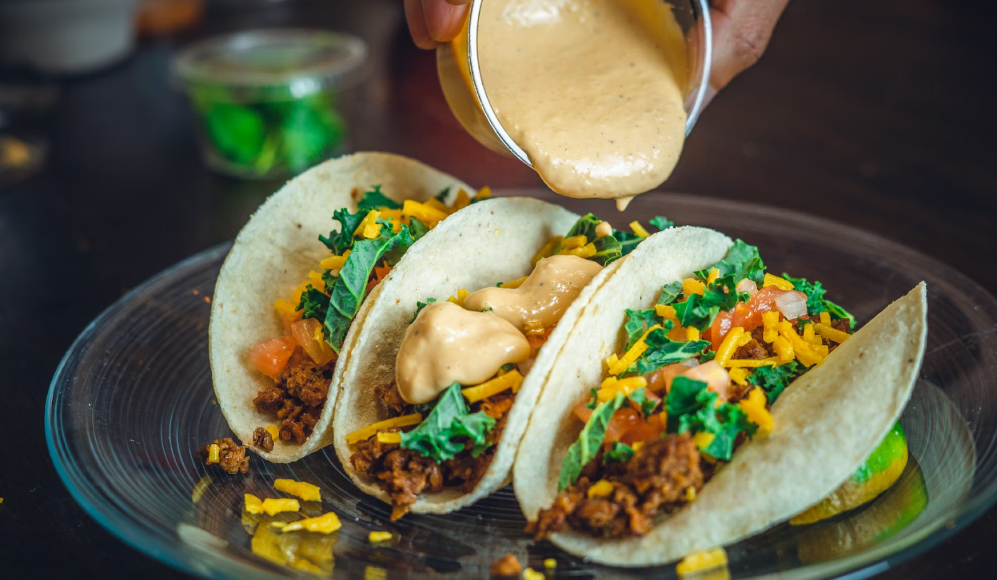 A hand is pouring a creamy sauce over three tacos filled with greens, cheese, and meat on a glass plate.