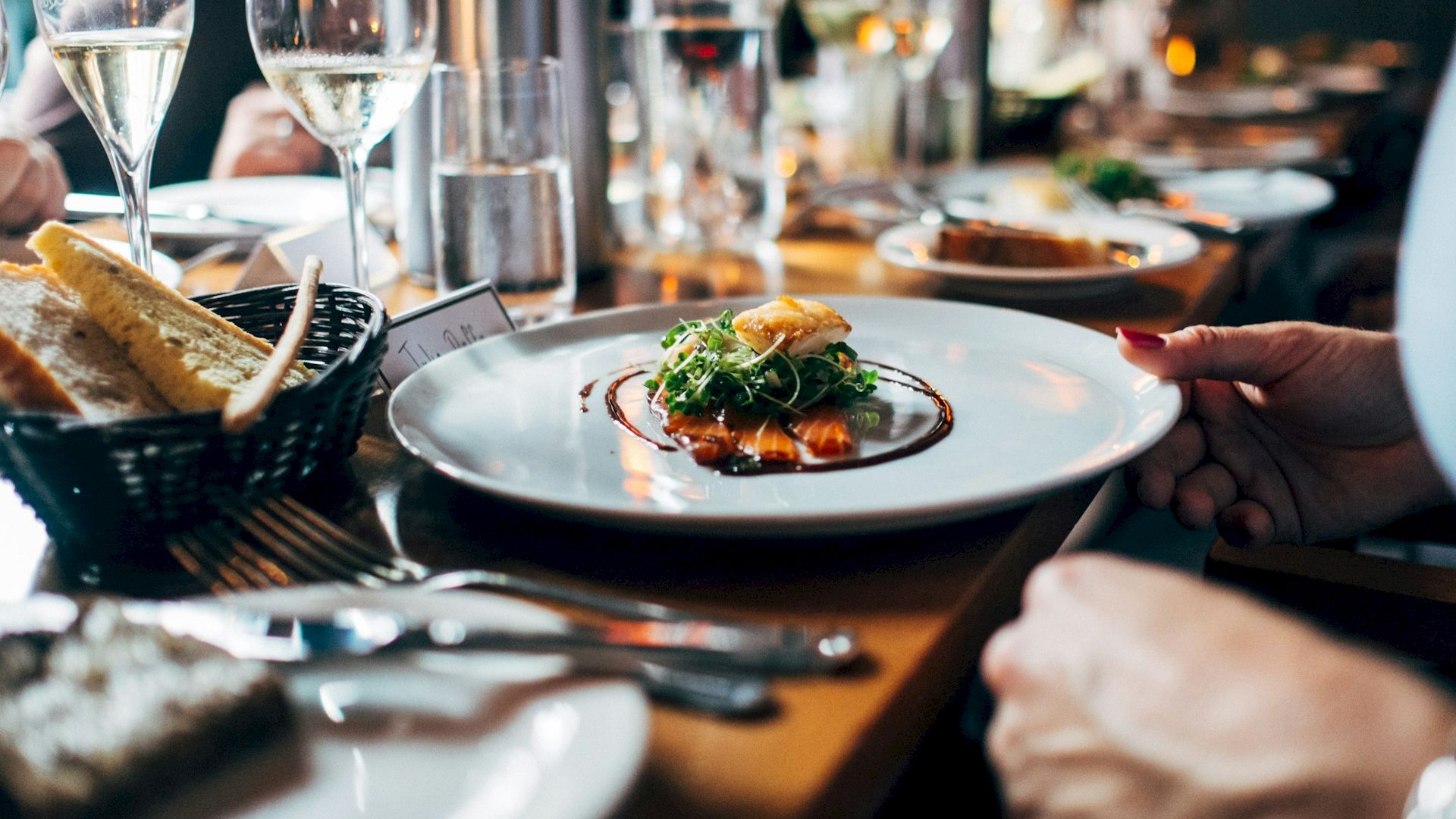 A gourmet dish with greens and sauce on a white plate is the focus; bread, wine glasses, and utensils are also on the wooden table.