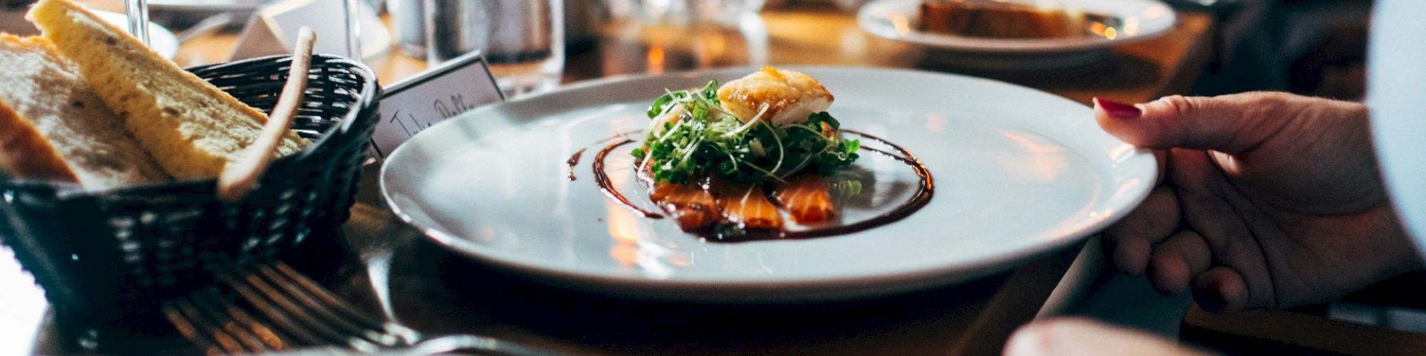 A gourmet dish with greens and sauce on a white plate is the focus; bread, wine glasses, and utensils are also on the wooden table.