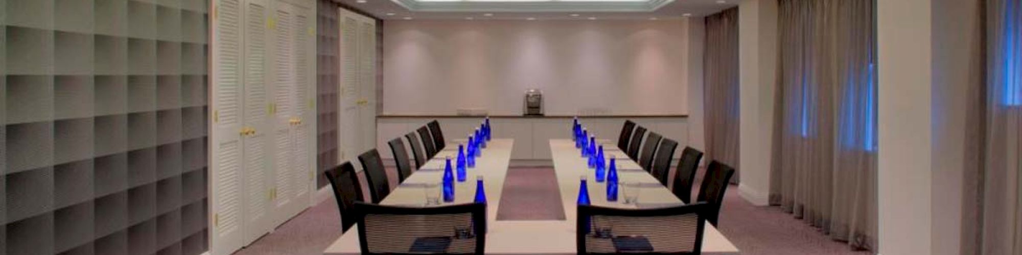 A modern conference room with a long table, chairs, and blue water bottles. The room has a patterned carpet and large windows with curtains.