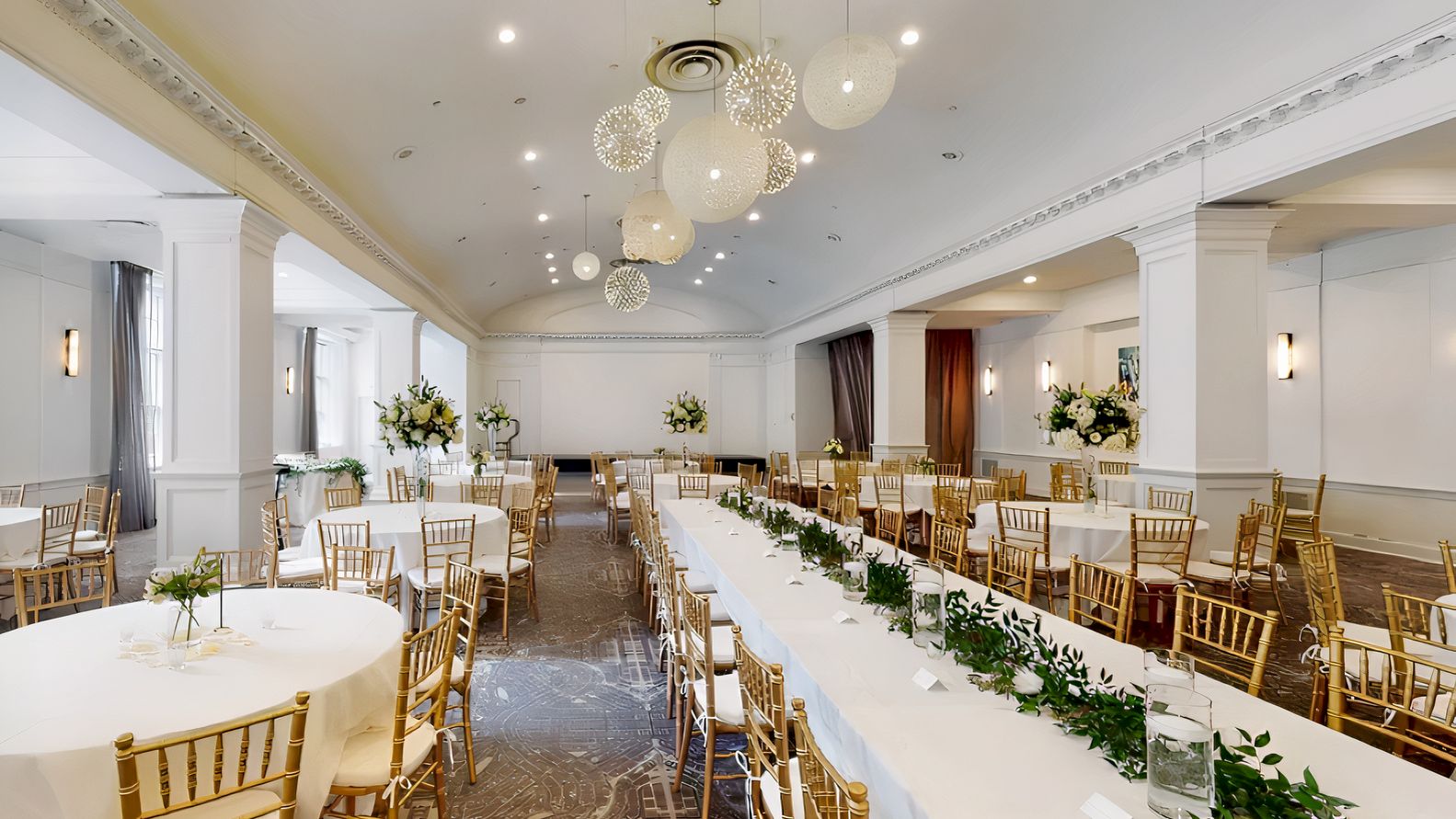 The image shows a decorated banquet hall with round and rectangular tables, gold chairs, floral arrangements, and overhead spherical lights.