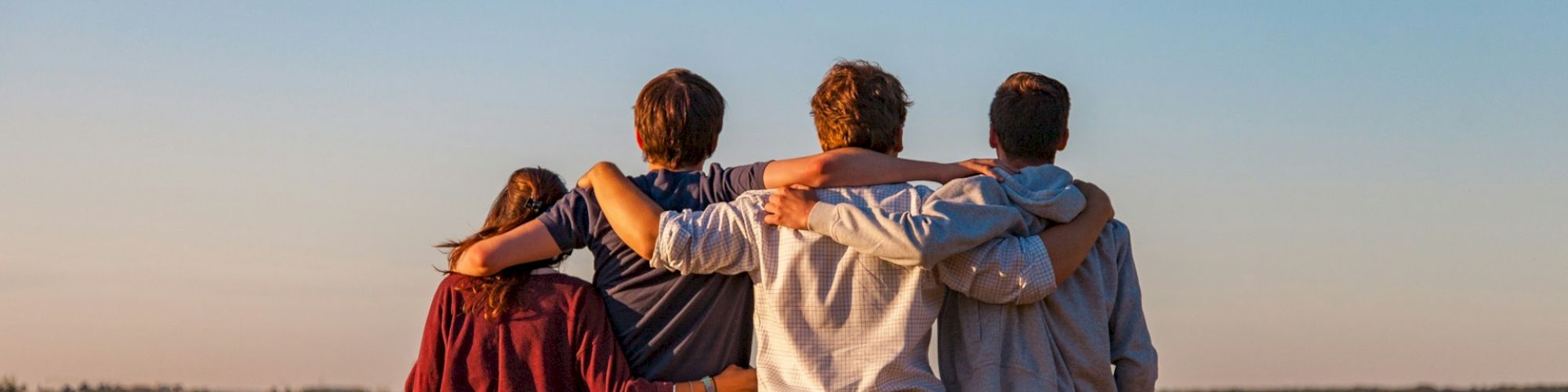 Four people standing in a golden field with their arms around each other, facing away from the camera, under a clear blue sky.