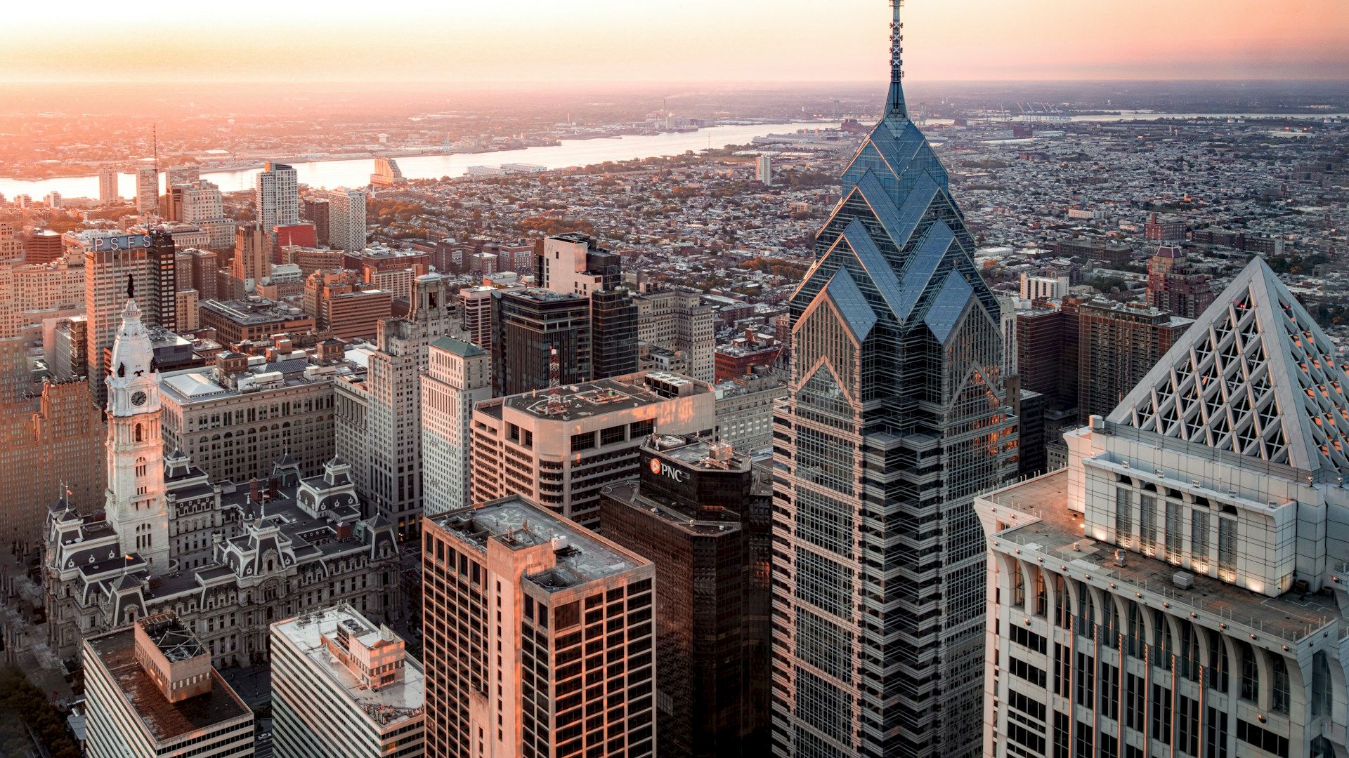 A cityscape at sunset with prominent skyscrapers, including a tall pointed building, and a river visible in the background, ending the sentence.
