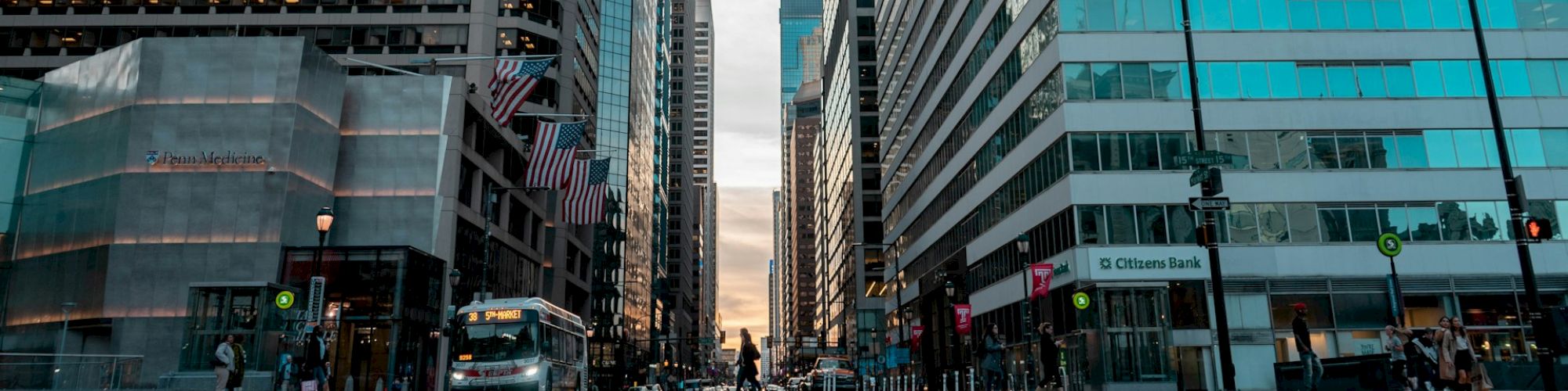 A city street flanked by tall buildings with a bus, streetlights, traffic signals, and pedestrians; the sky is visible between the skyscrapers.