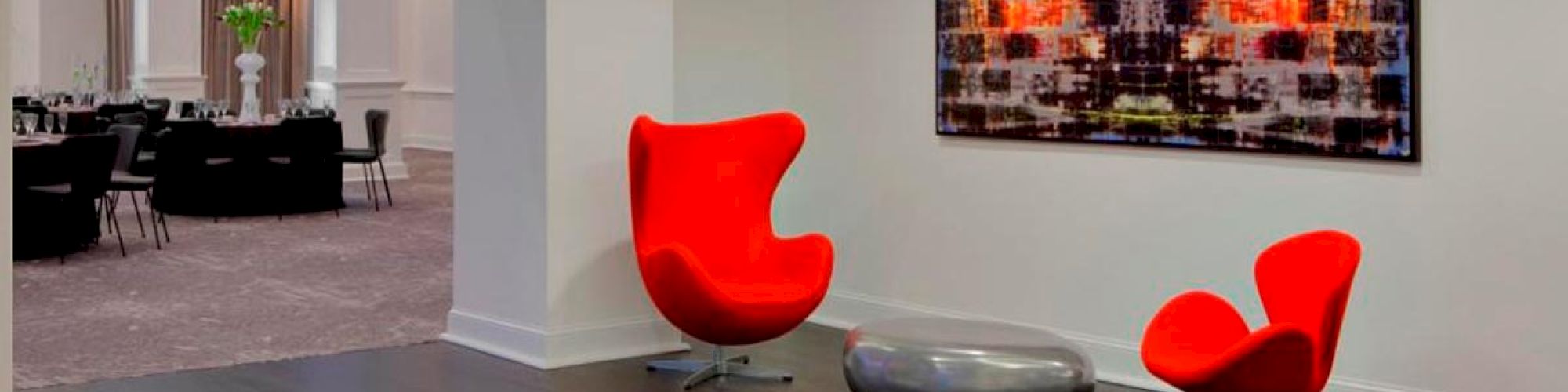 This image shows a modern lobby area with two red chairs, a sleek coffee table, and a colorful abstract painting on the wall under a circular ceiling light.