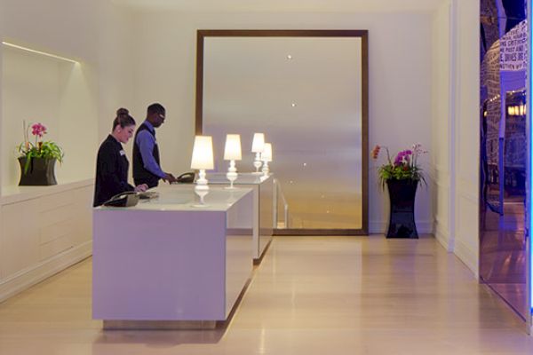 The image features a modern lobby with two people at a reception desk; the room has sleek furniture, potted plants, illuminated decorations, and a contemporary ambiance.