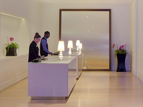 The image features a modern lobby with two people at a reception desk; the room has sleek furniture, potted plants, illuminated decorations, and a contemporary ambiance.
