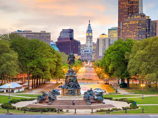 This image shows a city skyline with a prominent statue in the foreground, surrounded by trees and a park, under a colorful sky at sunset.