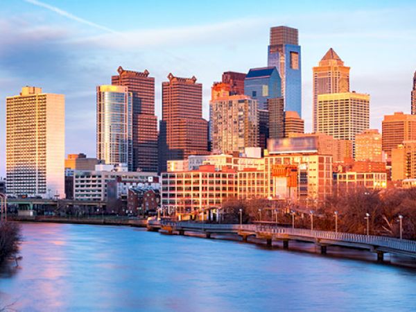 This image shows a city skyline at sunset, with tall buildings reflecting the sunlight, a river in the foreground, and a bridge crossing over the water.