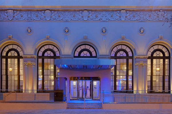 An illuminated building facade with arched windows, decorative detailing, and trees on each side, viewed during the evening or night.