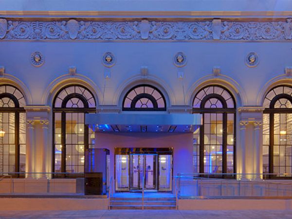 An illuminated building facade with arched windows, decorative detailing, and trees on each side, viewed during the evening or night.