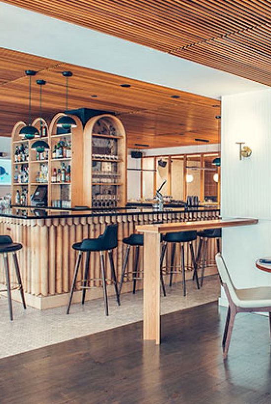 The image shows a modern bar area with high stools and tables, wooden ceiling, and a seating area with round tables and chairs in a spacious setting.