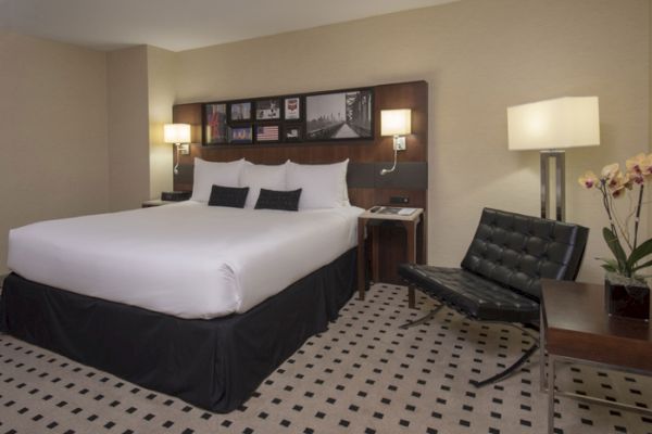 A modern hotel room features a large bed, a black chair, bedside tables with lamps, and a small table with a potted orchid.