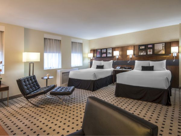 A hotel room with two double beds, modern furnishings, two chairs, a table, lamps, and a patterned carpet, lit by natural and artificial light.