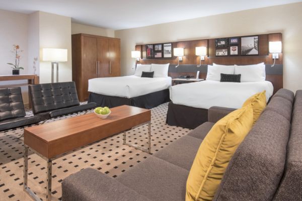 The image shows a spacious hotel room with two double beds, a sofa with yellow cushions, a wooden coffee table, and modern decor.