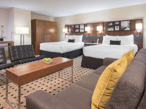 The image shows a spacious hotel room with two double beds, a sofa with yellow cushions, a wooden coffee table, and modern decor.