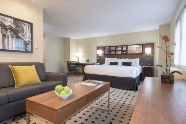 A modern hotel room features a large bed, sofa, coffee table with apples, desk, and artwork. Bright and well-lit with tasteful decor.