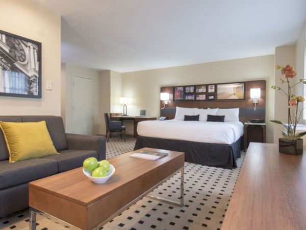 A modern hotel room features a large bed, sofa, coffee table with apples, desk, and artwork. Bright and well-lit with tasteful decor.