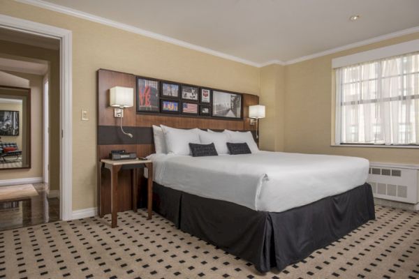 A hotel room features a large bed with white linens, two bedside tables with lamps, wall art, a window, and a hallway leading to the entrance.