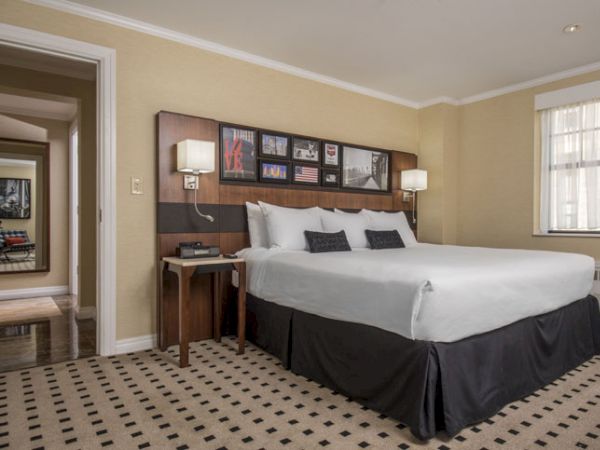 A hotel room features a large bed with white linens, two bedside tables with lamps, wall art, a window, and a hallway leading to the entrance.