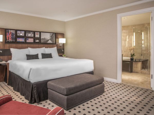 A modern hotel room with a large bed, bedside tables, lamps, a bench, and artwork. An open door reveals a bathroom with a tub.
