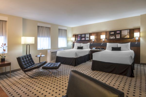 The image shows a modern hotel room with two double beds, a patterned carpet, lounge chairs, lamps, and framed artwork on the wall.