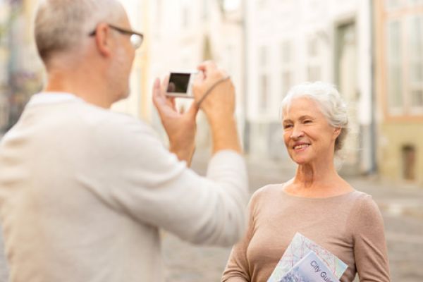 An older woman is posing for a photo being taken by an older man in an outdoor setting, holding a city guide.