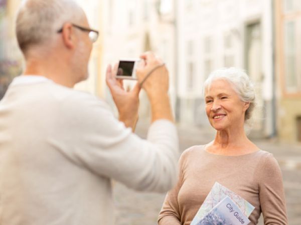An older woman is posing for a photo being taken by an older man in an outdoor setting, holding a city guide.