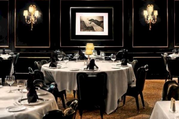 A dimly lit, elegant restaurant with round tables draped in white cloth, adorned with black napkins, wine glasses, and a single lit lamp on one table.