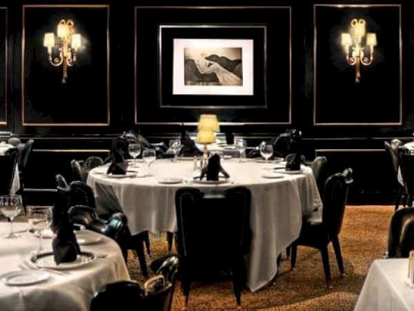A dimly lit, elegant restaurant with round tables draped in white cloth, adorned with black napkins, wine glasses, and a single lit lamp on one table.