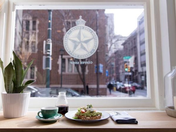 A plant, cup, and plate with food on a table by a window, showing a city street outside and a logo on the glass.
