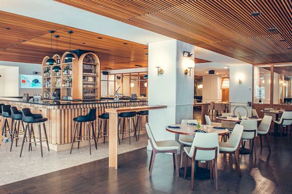 The image shows a modern, stylish bar area with high stools around the bar and several dining tables with chairs in an elegantly designed interior.